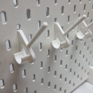 Puede incluir: Un panel perforado blanco con varios ganchos blancos insertados. El panel perforado tiene una cuadrícula de agujeros oblongos espaciados uniformemente. Los ganchos están diseñados para sujetar objetos y están angulados para brindar soporte. El diseño es simple y funcional.