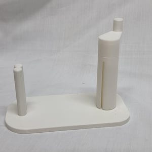 Pode incluir: Um suporte de bobina branco impresso em 3D. O suporte tem uma base retangular com dois postes verticais de um lado e um poste cilíndrico mais alto com uma pequena extensão angular do outro. O design é minimalista e funcional.