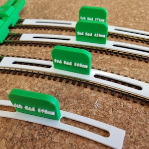 May include: Green and white model train track pieces with text indicating radius measurements. The tracks are set on a brown surface. The text on the green pieces reads "1st Rad 371mm", "2nd Rad 458mm", "3rd Rad 505mm", and "4th Rad 648mm".