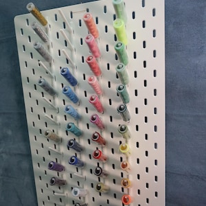Puede incluir: Un tablero perforado rectangular blanco con filas verticales de clavijas que sostienen varios carretes de hilo en un espectro de colores que incluye oro, rosa, azul, verde y naranja. El tablero tiene una cuadrícula de ranuras espaciadas.
