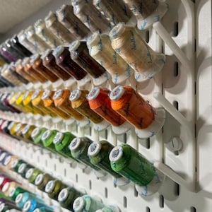 May include: A white pegboard with rows of colorful thread spools organized in clear plastic holders. The spools are labeled "Sulky" and are arranged by color.