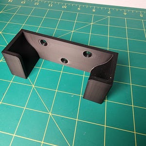 May include: A black plastic wall mount with three holes for mounting screws. The mount has a curved shape to hold an object.