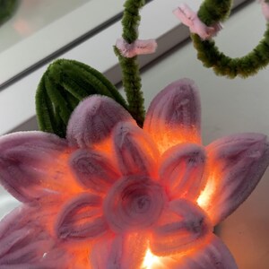 Op de afbeelding: Een handgemaakte bloem van pluizige pijpenragers met lichtroze bloemblaadjes en een groene steel. Het midden van de bloem gloeit met een warm oranje licht en de steel is voorzien van kleine roze accenten en een gekruld bovendeel met groene bladeren.