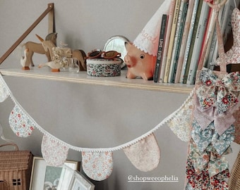 May include: A decorative shelf displays a variety of items, including books, a piggy bank, and a floral garland. A small pink jewelry box and framed photos sit on the surface below. The garland features floral fabric pennants.