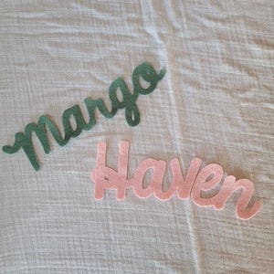May include: Two felt name cutouts, "Margo" in green and "Haven" in pink, displayed on a textured, light-colored surface. The cursive lettering is a decorative element, suitable for personalized crafts or room decor.