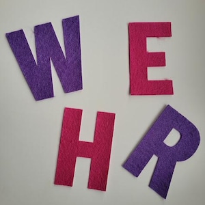 Op de afbeelding: Vier individuele viltletters gerangschikt om het woord WEHR te spellen. De letters W, H en R zijn paars, terwijl de letter E een levendige roze kleur heeft. Elke letter heeft een matte textuur en een strakke bloklettervorm uit vilt.
