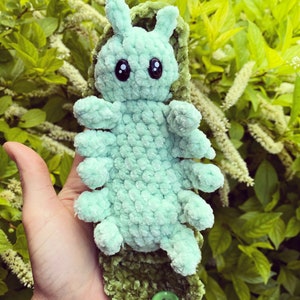 Op de afbeelding: Een lichtgroen gehaakt knuffeltje in de vorm van een rups met zwarte ogen en een groen gehaakt zakje in de vorm van een blad met een knoop.
