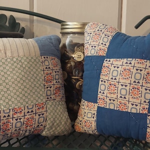 May include: Two small, square patchwork pillows with a vintage aesthetic. One pillow features a mix of blue, white, and patterned fabrics, while the other showcases a blue and floral design. The pillows are placed on a green metal surface.