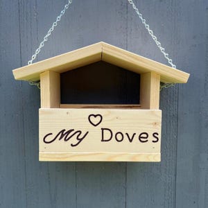 May include: A wooden birdhouse with a pitched roof and a rectangular opening. The words "My Doves" are written on the front, with a heart symbol above the word "Doves". The birdhouse is suspended by a silver chain.