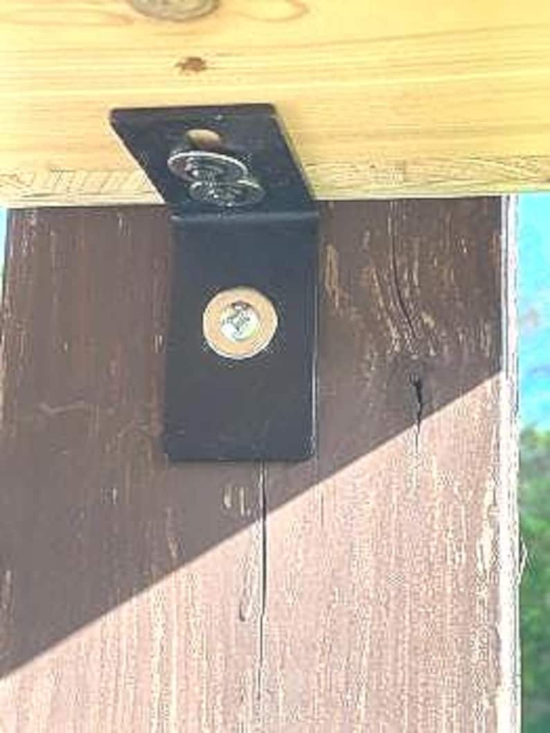 May include: Close-up of a black metal bracket attached to a wooden beam with two screws. The bracket has a rounded edge and a flat surface.