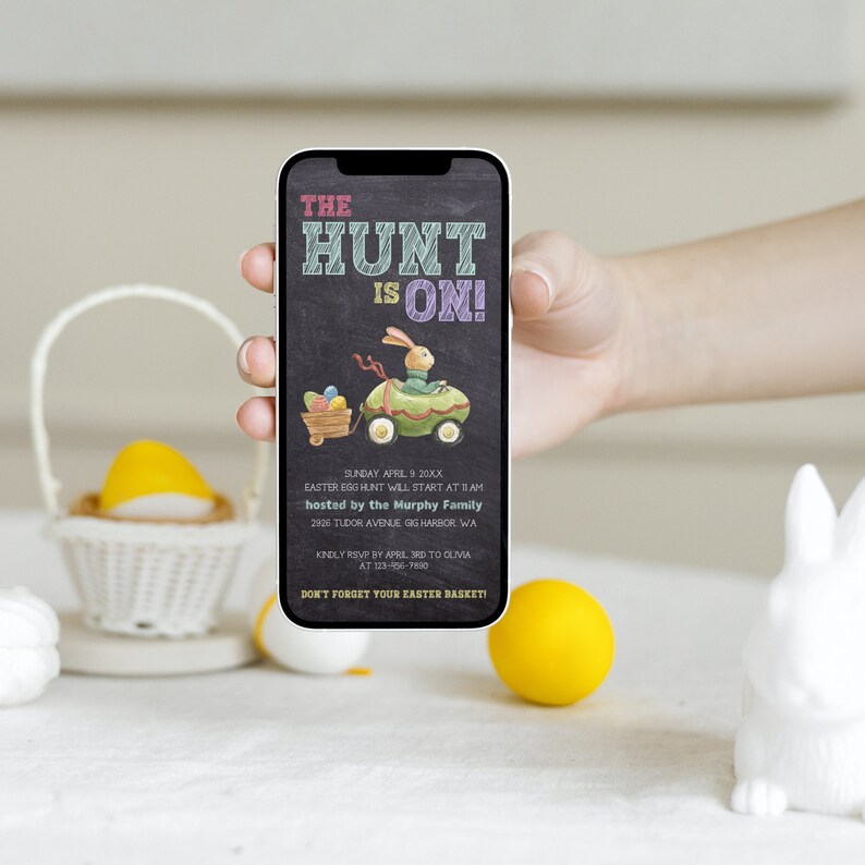 May include: A smartphone displaying an invitation to an Easter egg hunt. The invitation is on a black chalkboard background with colourful chalk writing. The text reads "The Hunt is On!" and includes details about the event, including the date, time, location, and host. A cartoon bunny is driving a green car with a basket of eggs.