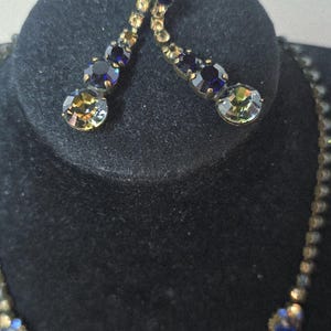 May include: A gold-coloured jewellery set comprising a necklace and earrings. The necklace and earrings are decorated with blue and iridescent gemstones. The earrings have a dangling design. The jewellery is displayed on a black velvet surface.