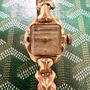 May include: A vintage gold-toned wristwatch with a square face. The watch face is marked "Wittnauer" and features gold hands and hour markers. The watch has an ornate, textured band and a crown on the side. The background is a green and white patterned surface.