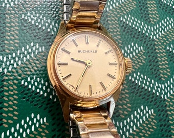 May include: A gold-toned wristwatch with a round face and a gold metal band. The watch face has Roman numeral hour markers and the word "BUCHERER" printed on it.