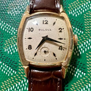 Peut inclure: Une montre-bracelet Bulova vintage avec un boîtier rectangulaire doré et un bracelet en cuir marron. Le cadran de la montre est de couleur crème avec des chiffres et des aiguilles noirs. La montre est posée sur un fond vert à motifs.