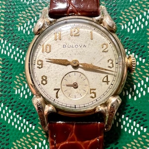 Op de afbeelding: Vintage Bulova polshorloge met een goudkleurige kast en een crèmekleurige wijzerplaat. Het horloge heeft gouden wijzers en cijfers, een kleine secondenwijzer en een bruine leren band. De wijzerplaat toont de merknaam Bulova.