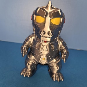 May include: A metallic silver and black figurine of a robotic dinosaur with yellow eyes. The figure has a detailed design with rivets and panels, and is posed in a standing position. The toy is a collectible item.
