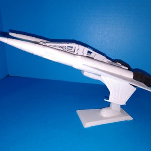 May include: A white model airplane with a sleek design, featuring a long, pointed nose and wings. The model is mounted on a rectangular base. The airplane is set against a bright blue background.