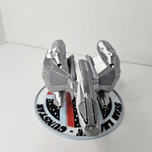 Könnte beinhalten: Ein silbernes 3D-gedrucktes Modell eines Raumschiffs mit vier Triebwerken. Das Raumschiff steht auf einem runden Sockel mit der Aufschrift "Sun Lair - Gunstil".