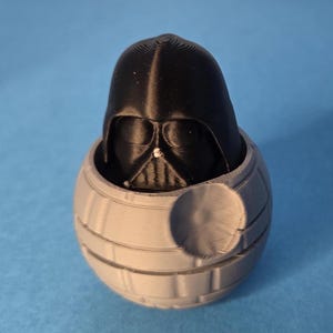 May include: A 3D-printed Darth Vader head is housed within a grey Death Star model. The black Vader helmet is detailed, and the Death Star has a textured surface. The background is a solid light blue.