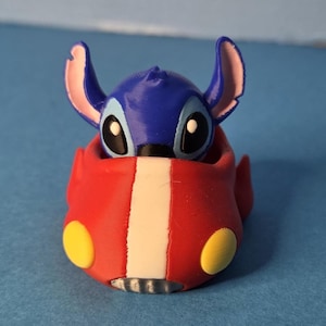 May include: A 3D-printed figurine of the character Stitch from the Disney film Lilo & Stitch. The blue Stitch is inside a red vehicle with yellow accents and a white stripe. The figurine is on a blue surface.