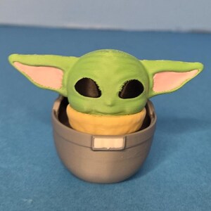 May include: A green figurine of a character with large ears and black eyes, seated in a gray pod. The character's ears have pink inner details, and it has a yellow head. The pod has a small, rectangular white detail.