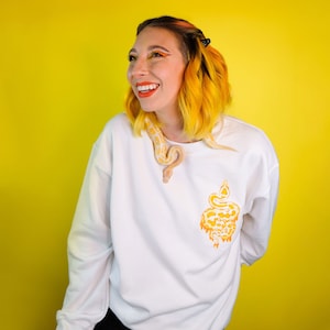 Könnte beinhalten: Ein weißer Pullover mit einem gelben Schlangendesign auf der Vorderseite. Die Schlange ist um den Hals der Person gewickelt, die den Pullover trägt. Die Person hat gelbes Haar und lächelt.