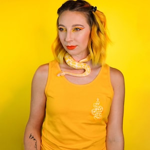 Könnte beinhalten: Eine Frau mit gelben Haaren trägt ein gelbes Tanktop mit einem Schlangenmotiv auf der Vorderseite. Sie trägt eine weiße Schlange um den Hals. Der Hintergrund ist gelb.