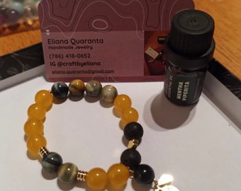 May include: A yellow and tiger's eye gemstone bracelet with a gold charm and lava beads. The bracelet is next to a business card and a bottle of essential oil labeled "MENTHA PIPERITA".