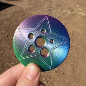 Puede incluir: Un disco metálico circular con un diseño de estrella de cinco puntas y múltiples recortes circulares. El disco tiene un acabado iridiscente arcoíris.