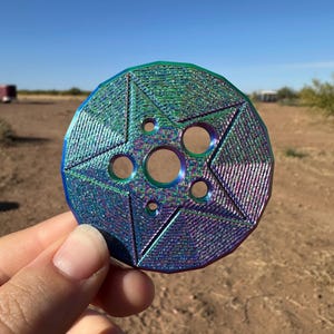 May include: A multi-colored, iridescent metal object with a star-like design. The object has a series of circular holes and geometric patterns. The color shifts between blue, green, and purple. The object is held in a hand against a natural outdoor background.