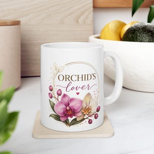 May include: White ceramic mug featuring the text "ORCHID'S lover" in elegant script, surrounded by a floral design with pink and cream orchids. The mug sits on a beige coaster, with a handle on the right side.
