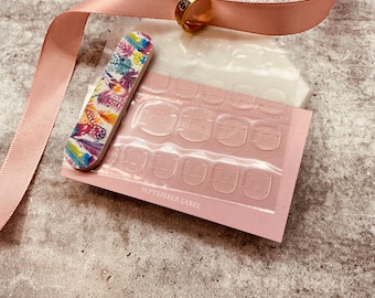 Puede incluir: Un paquete rosa y blanco que contiene pestañas adhesivas transparentes para uñas. El paquete también incluye una lima de uñas con un diseño de plumas de colores. El texto "SEPTEMBER LABEL" está impreso en el paquete.