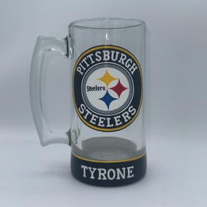 May include: A clear glass beer mug with a handle. The mug features the Pittsburgh Steelers logo in the center, with a black and gold base that reads "TYRONE" in white lettering. The logo includes a yellow star, a red star, and a blue star.