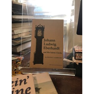 May include: A brown book with a black and white illustration of a tall clock on the cover. The title of the book is "Johann Ludwig Eberhardt and His Salem Clocks" by Frank P. Albright. The book is part of "The Old Salem Series."