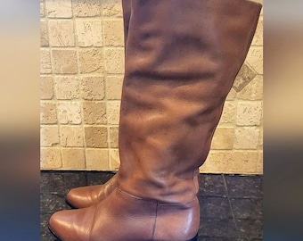 May include: A pair of brown leather knee-high boots with a low heel. The boots are made of a soft, supple leather and have a slightly slouched look.