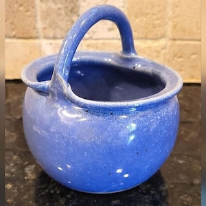 May include: A small, blue ceramic bowl with a handle. The bowl has a speckled glaze and a rounded shape.