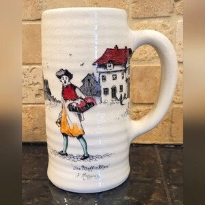 May include: White ceramic mug with a hand-painted scene of a man in a red and yellow outfit carrying a basket of muffins. The scene is set in a European town with a red-roofed building. The mug has the text "The Muffin Man" and a signature at the bottom.
