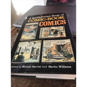 May include: A Smithsonian Book of Comic-Book Comics book cover with a black background and four comic book panels featuring illustrations of characters from different comic strips. The book title is in white text with a red outline. The book cover also includes the text "Edited by Michael Barrier and Martin Williams".