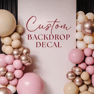 May include: A white backdrop with the text "Custom BACKDROP DECAL" in burgundy script. The backdrop is decorated with clusters of pink, beige, and metallic balloons, creating a festive and celebratory atmosphere.