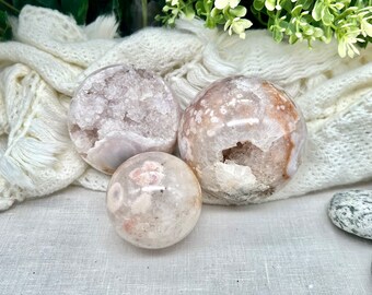 Puede incluir: Tres esferas de ágata rosa y blanca con un interior cristalizado. Las esferas están sobre una superficie de tela blanca.