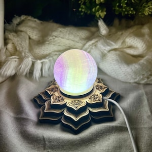 Puede incluir: Un orbe iridiscente brillante se asienta sobre una base de madera con un diseño de mandala decorativo. La base tiene un cable conectado a ella, lo que sugiere que es una luz o una lámpara.