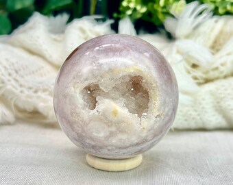 Puede incluir: Una esfera de ágata blanca y rosa con una gran abertura de geoda de cristal natural. La esfera está sentada sobre un soporte de madera.