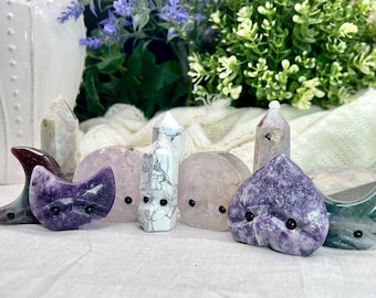 Puede incluir: Una colección de siete pequeñas figuritas de cristal talladas en varias formas, incluyendo un corazón, una luna creciente y una torre. Cada figurita tiene ojos negros y está hecha de un tipo diferente de cristal, incluyendo amatista, cuarzo rosa y cuarzo claro.