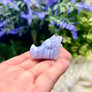 May include: A blue agate dragon sculpture carved from a blue and white banded agate stone. The dragon is facing to the right and has a detailed face and wings.