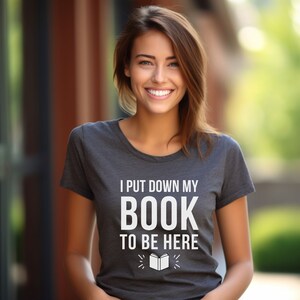 Puede incluir: Una camiseta gris con el texto "Dejé mi libro para estar aquí" y una pequeña ilustración de un libro abierto.