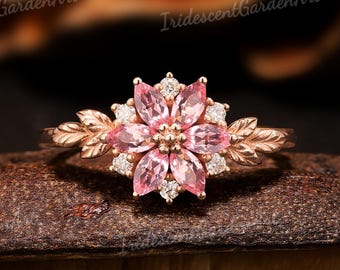 May include: A rose gold ring featuring a flower design with pink petals and small white accent stones. The ring has leaf-shaped details on the band. The ring is displayed on a brown surface.
