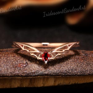 May include: A rose gold ring with a bat wing design. The ring features a pear-shaped, deep red gemstone at the center. The wings extend outward from the gemstone, creating a unique and elegant look. The ring is displayed on a dark surface.