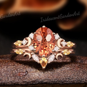 Op de afbeelding: Een roségouden verlovingsring met een ovaal geslepen, perzikgekleurde edelsteen als middensteen. De ring is geaccentueerd met kleinere witte en gele edelstenen en halve maan ontwerpen. De ring wordt gepresenteerd op een donkere, getextureerde ondergrond.