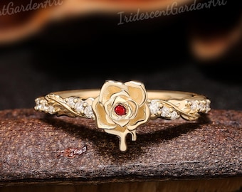 May include: A gold ring featuring a central rose-shaped motif with a small red stone at its center. The band is designed with a vine-like texture and is accented with small, clear, sparkling stones along the sides of the rose centerpiece.